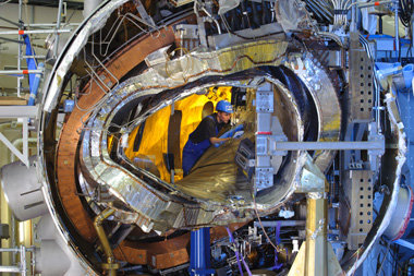Ende 2011 war das Innenleben von Wendelstein 7-X noch sichtbar; jetzt ist die ringförmige Anlage geschlossen. Vom Zentrum nach außen: das Plasmagefäß, eine der verwundenen Stellaratorspulen (silberfarben), eine ebene Spule (kupferfarben), die Stützstruktur (rechts) und das Außengefäß zusammen mit zahlreichen Kühlleitungen und Stromverteilern.