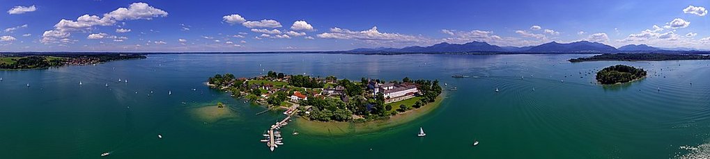 Panorama einer Insel im klaren blauen See, gesäumt von Segelbooten, mit einer Bergkette am Horizont unter einem leicht bewölkten Himmel.
